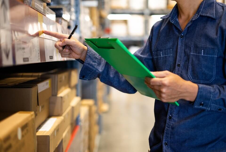 counting inventory in a warehouse using a clipboard while they are on shelves in cardboard boxes packed up until they are shipped and delivered